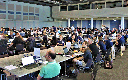 Photograph of a Connectathon event, showing many people sharing long desks, which span multiple rows. Each person is an engineer working for a vendor, which is connected to a network and is testing their software with that of other competitors, to ensure proper interoperability.